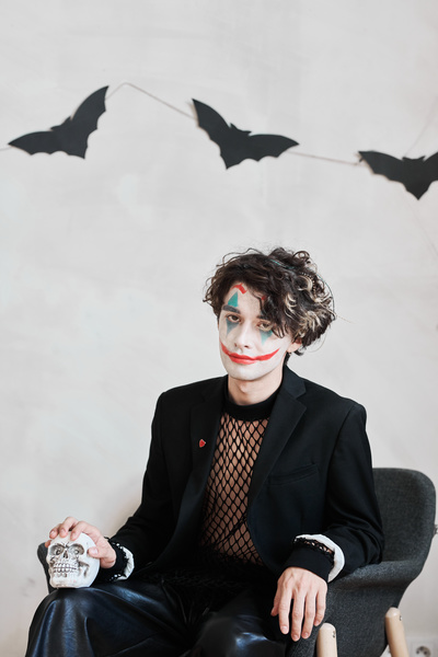 Man with Joker Makeup Is Sitting against Background of Wall with Bats with Skull in His Hand